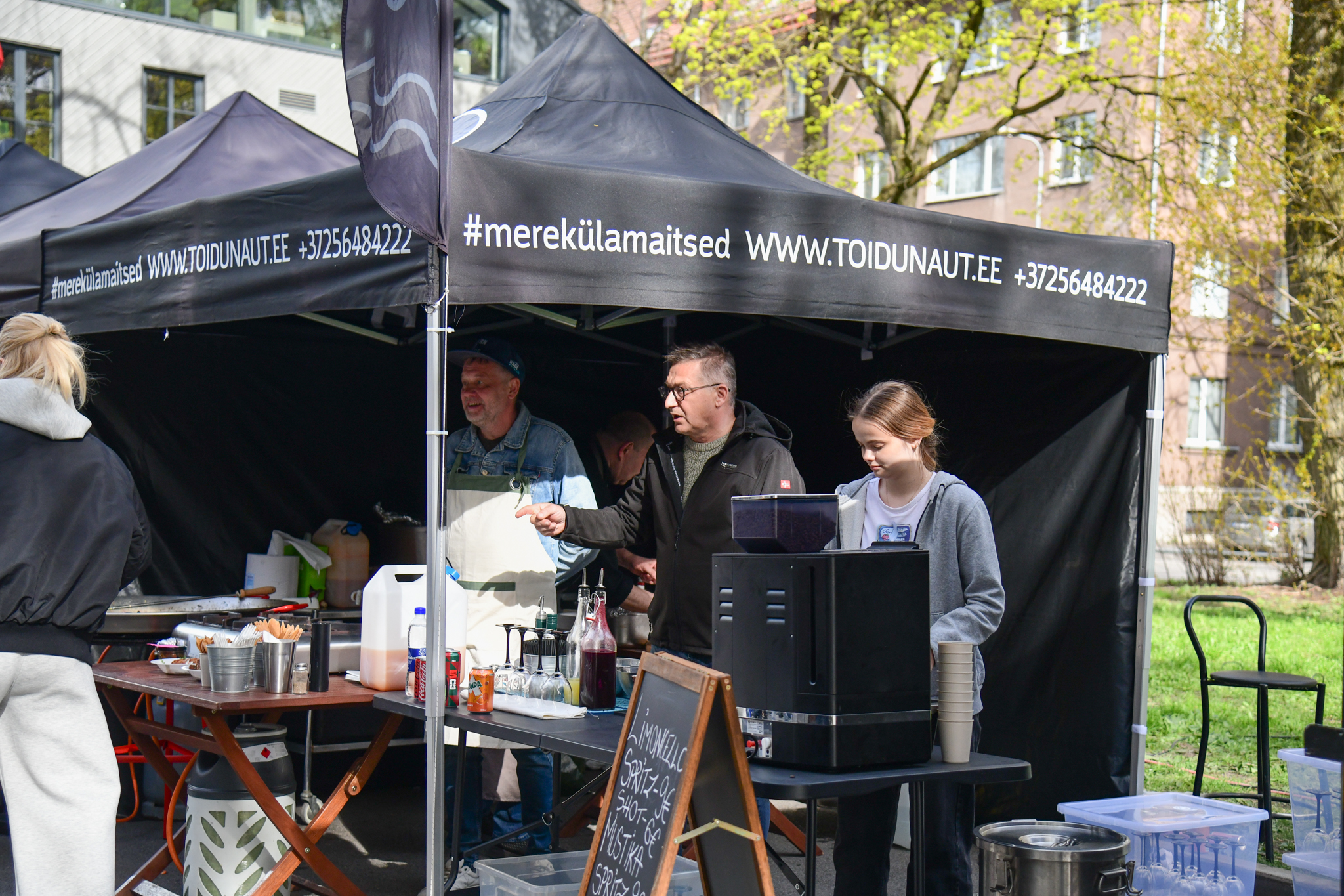 Outdoor market stall with a black canopy and chalkboard menu; people serving drinks under the tent, urban buildings in background.